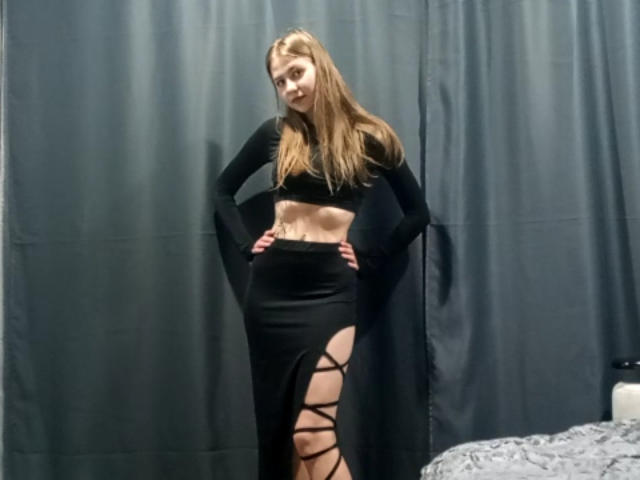 Margret Warriner está em pé, com as mãos nos quadris, vestindo uma blusa cropped preta de manga comprida e uma saia preta com grandes recortes laterais que revelam suas pernas e nádegas..