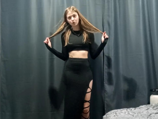 MargretWarriner with long blonde hair, wearing a black crop top and high-waisted skirt with lace-up sides, holding her hair, standing in front of dark curtains.