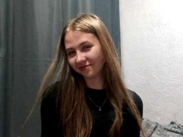 MargretWarriner has long, straight blonde hair, a black top, and a subtle smile, standing in a room with a white wall and grey curtain. She's wearing a necklace.