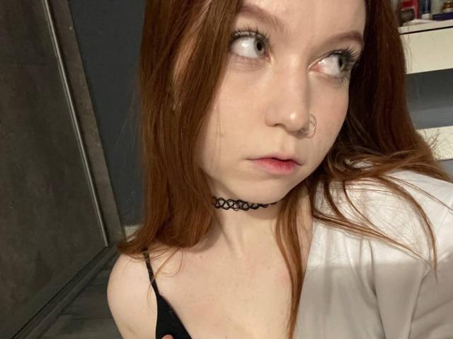 LilliamChevalier with red hair, nose ring, and black choker, looking to the side, wearing a white shirt and black strap, with her tits barely visible.