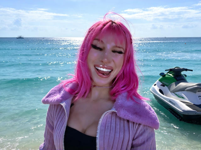 Madiy69 with pink hair, smiling with tongue out, wearing a purple zip-up jacket and black top, standing in clear water, green and white jet ski in background.