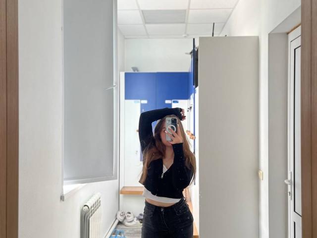 Dorthy Kuzma met lang bruin haar, een zwarte top, een witte cropped top, zwarte broek met hoge taille, die haar arm omhoog steekt, een spiegel-selfie maakt, met een zichtbare telefoon, in een witte gang met blauwe kasten..