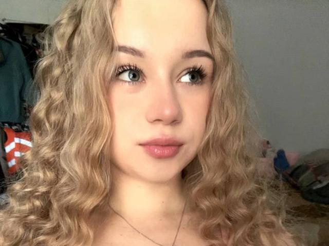 Dorthy Kuzma tem cabelos loiros encaracolados, olhos azuis e lábios rosados. Ela está olhando para o lado com um leve sorriso, usando um colar fino. Seus seios não estão visíveis..