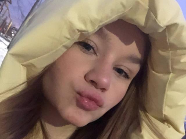 Het gezicht van DorthyKuzma, met een pluizige gele capuchon, lichtjes opgetrokken roze lippen, bruine ogen en lang bruin haar. Ze draagt een pluizige gele jas en ziet er schattig en ontspannen uit..