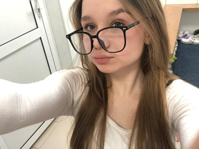 DorthyKuzma with long brown hair, wearing black-framed glasses and a white long-sleeve shirt, pouting her pink lips, taking a selfie, showing her blue eyes and fair skin.