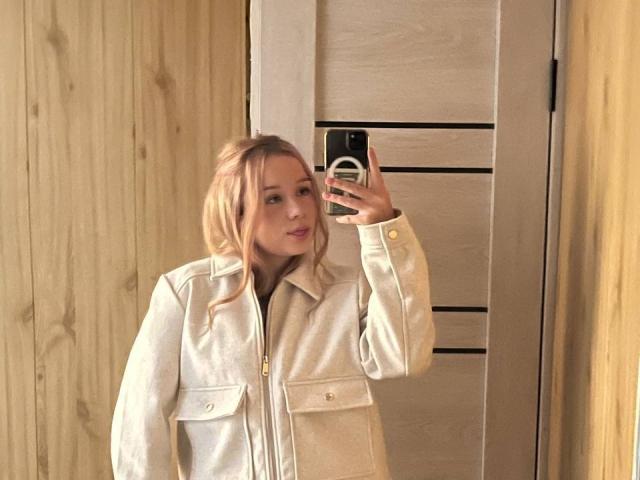 DorthyKuzma with blonde hair, wearing a beige zip-up jacket, taking a mirror selfie with her phone, showing a slight smile, in a wooden room.