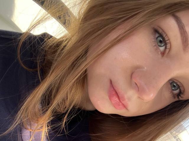 Le visage de Dorthy Kuzma, ses yeux bleus, ses lèvres roses, ses cheveux châtains clairs, un léger froncement de sourcils, elle porte un haut sombre, la lumière du soleil sur sa joue, des taches de rousseur visibles, une peau douce, des cheveux légèrement ondulés..