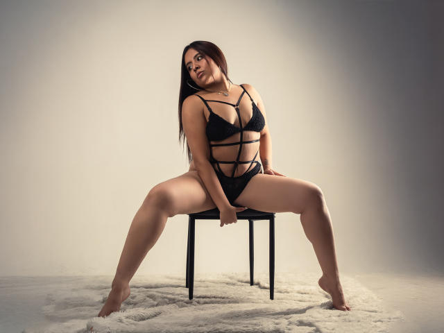 GabbyRondon está sentada em uma cadeira, com as pernas abertas, vestindo lingerie preta com alças finas, expondo sua vulva. Ela tem cabelos castanhos longos, um colar e uma expressão confiante e sexy..