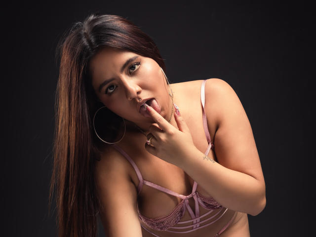 GabbyRondon, avec de longs cheveux bruns, un soutien-gorge en dentelle rose et des boucles d'oreilles cerceaux, suce son doigt, la langue dehors, sur un fond sombre. Seins, poitrine, et une bouche légèrement ouverte..