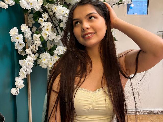 TrishMuhlbauer with long brown hair, smiling, wearing a yellow strapless top, hand in hair, white flowers in background, teal curtain, smiling at camera, cute, tan skin.