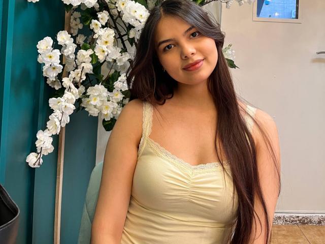 TrishMuhlbauer with long dark hair, yellow lace-trimmed tank top, and cute smile, standing next to white cherry blossoms, looking directly at the camera. Her tits are medium-sized.