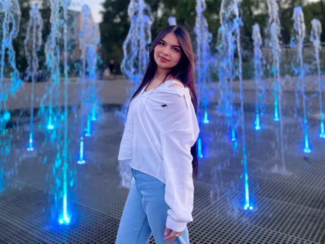 TrishMuhlbauer with long dark hair, white off-shoulder shirt, and light blue pants, standing in front of blue-lit water fountains, smiling slightly. Her ass is slightly visible.
