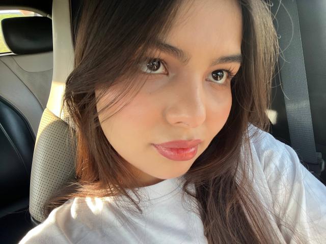 TrishMuhlbauer's face, showing brown eyes, pink lips, and straight brown hair, with a white shirt, sitting in a car's front seat. Her ass is out of frame.