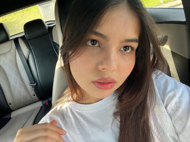 TrishMuhlbauer's face in a car, brown eyes, long dark hair, pink lips, white shirt, hand on chest, looking at camera, sunlight from window, black and grey car seat.