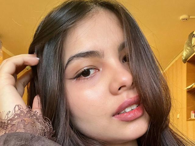 TrishMuhlbauer's face with straight, dark brown hair partially covering her right eye. She has light brown skin, subtle makeup, and pink lips. Her hand's in her hair.