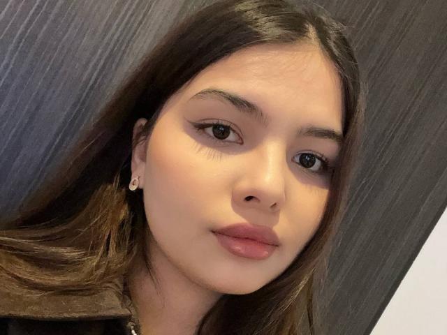 TrishMuhlbauer has smooth brown skin, long brown hair, and full pink lips. She's wearing a brown jacket with a small earring in her left ear.