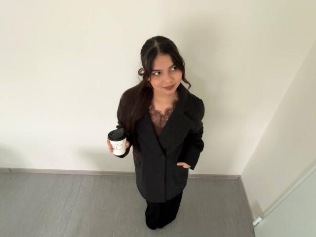 TrishMuhlbauer, dark-haired, wearing black suit with lace top, holding coffee cup, standing on gray floor, hand in pocket, looking up with slight smile.