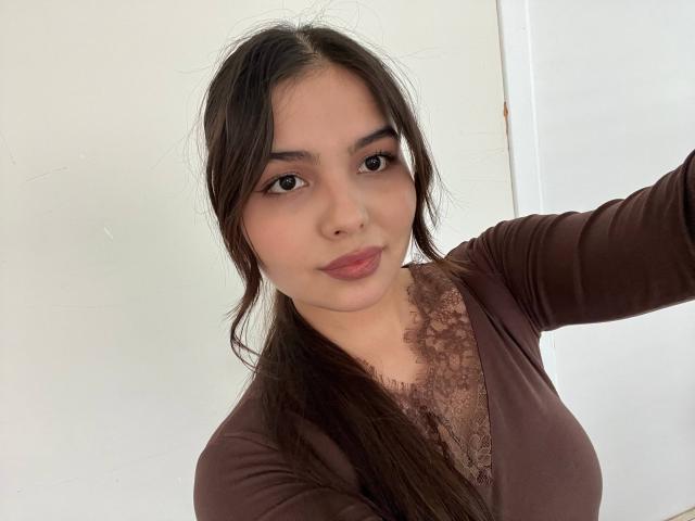 TrishMuhlbauer has long dark brown hair, brown eyes, and is wearing a brown lace-trimmed top. Her tits are small and she's taking a selfie with a slight smile.