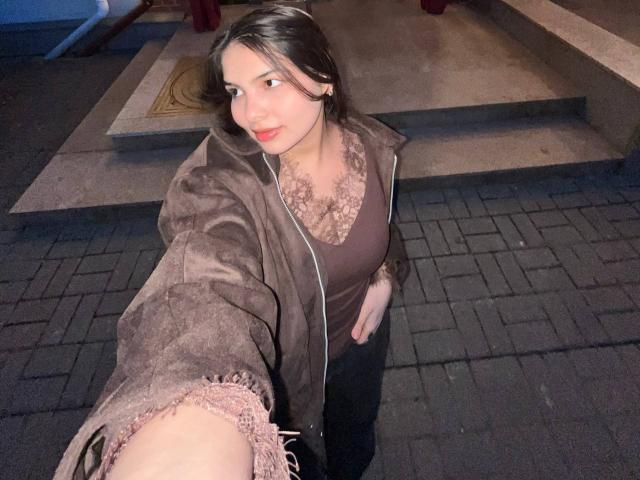 TrishMuhlbauer with brown hair, wearing a brown suede jacket, lace necklace, brown top, and black pants, taking a selfie, showing her slightly parted lips and light skin.