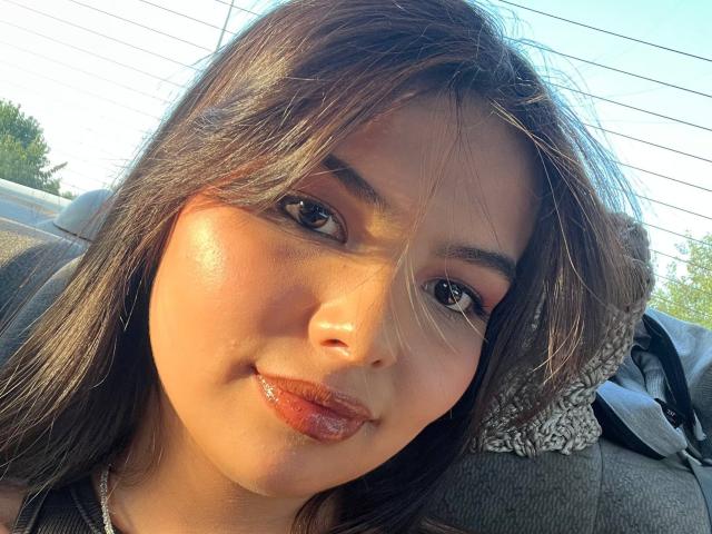 TrishMuhlbauer has glossy brown lips, dark brown eyes, and shiny, sunlit brown hair, with a subtle smile, looking at the camera in a car.