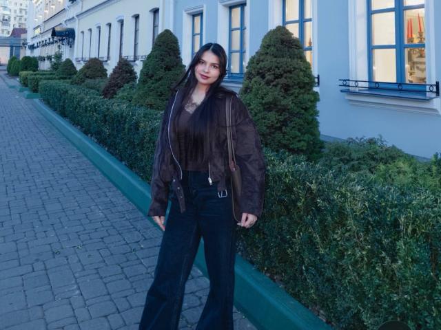 TrishMuhlbauer with long black hair, brown jacket, black top, black pants, standing on a brick sidewalk, green bushes, and white building with windows in the background.