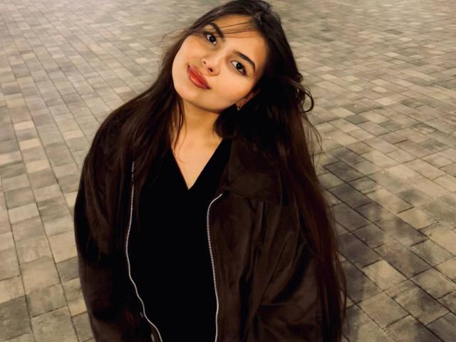 TrishMuhlbauer with long black hair, wearing a black zip-up jacket and black shirt, smiling slightly, standing on a tiled floor, looking up at the camera.