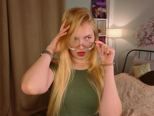 CarmelDelay with long blonde hair, green top, and glasses, making a peace sign with her eyes closed and red lips. She's holding her glasses with both hands.