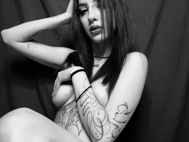 SimonneBrustkern, topless, with long dark hair, tattoos on arms, covering small tits with hand, wearing black bracelet, black choker, slightly parted lips, looking at camera, black and white.