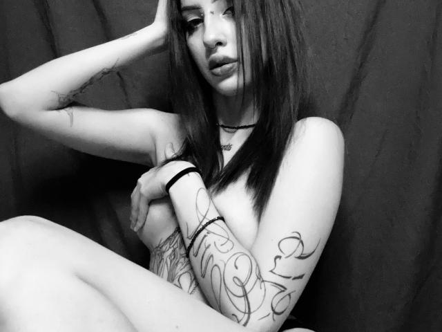 SimonneBrustkern with long dark hair, tattoos on arms and side, covering her tits with one arm, sitting against dark background, looking at camera with pouty lips, black bracelet.
