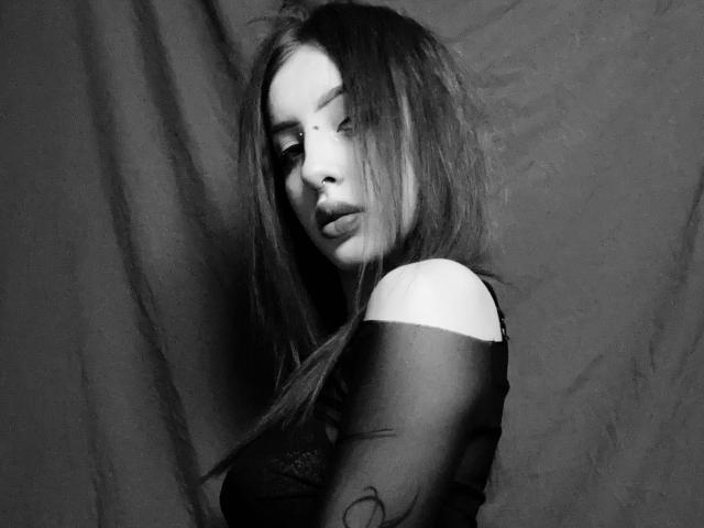 Black-and-white photo of SimonneBrustkern with long, messy hair, pouty lips, and a small beauty mark on her cheek, wearing a sheer, off-shoulder top, showing her tits.