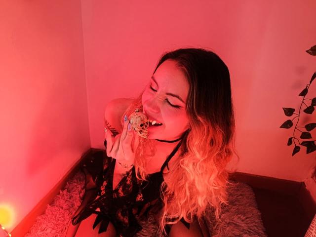 AlanisBlanc with long blonde to brown ombre hair, black lace lingerie, smiling, licking a skull-shaped lollipop, eyes closed, sitting on a furry rug, red-pink lighting.
