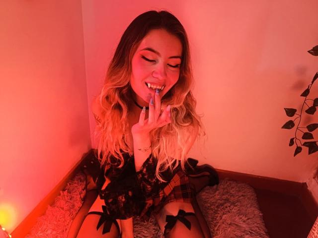 AlanisBlanc with blonde wavy hair, smiling, biting finger, wearing black lace and ribbons, sitting on a fluffy rug, red light, plant in corner, looking sexy.