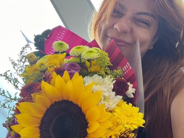 Samanta Carter hält einen farbenfrohen Blumenstrauß mit großen, gelben Sonnenblumen, grün-weißen Chrysanthemen und violetten Blumen. Sie lächelt mit geschlossenen Augen, hat braunes Haar und ein rosa Band..