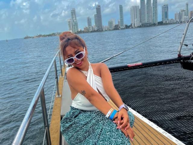 SamantaCarterr with brown hair in a bun, wearing white sunglasses, white halter top, blue patterned skirt, and bracelets, sitting on a boat, showing her tattooed arm.