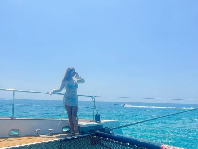 Samanta Carter in einem engen, weißen, langärmeligen Kleid, stehend auf einem Boot, mit ihrem Handy am Ohr, mit hellem, blauem Meer und klarem Himmel im Hintergrund..