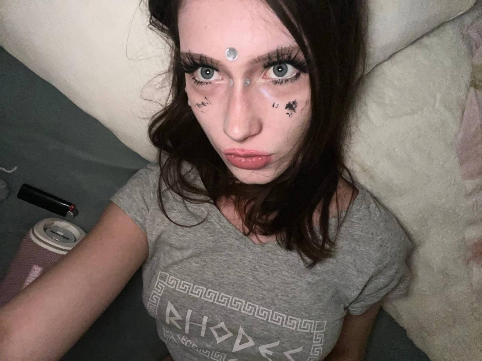 Danika Leitao, com cabelo castanho escuro, maquiagem facial branca, marcas de estrelas pretas e uma camiseta cinza, segurando uma caneca rosa com tampa preta, olhando para a câmera..
