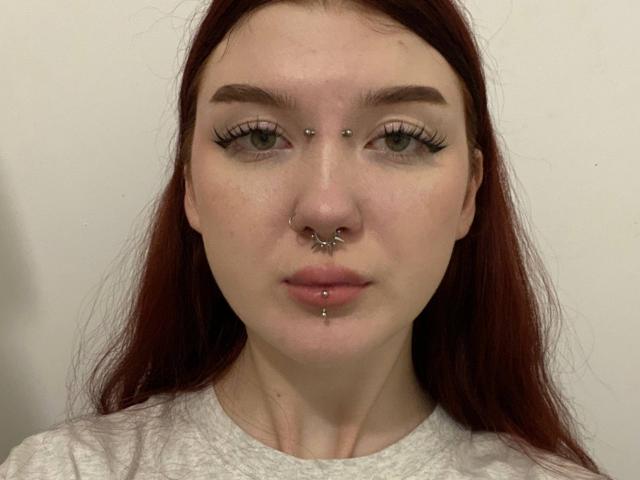 Johanne Kazmierczak tem cabelo ruivo, olhos verdes e várias perfurações faciais: no nariz, no septo e no lábio inferior. Ela está vestindo uma camisa branca e olhando diretamente para a câmera..