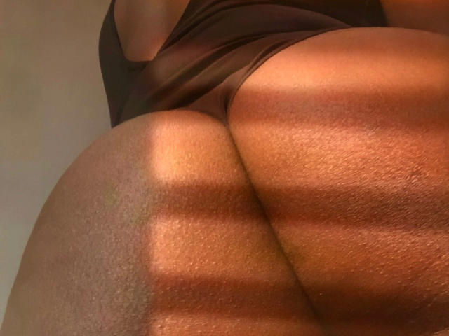SexyKayla-hot's ass, highlighted by warm sunlight, casts shadows on her dark brown, textured skin. Her black underwear slightly rides up, emphasizing her round, voluptuous buttocks.