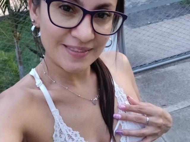 SolaraDubois, con gafas, sonriendo, sosteniendo su sujetador blanco de encaje, mostrando sus pequeños pechos, uñas rosadas y un anillo, al aire libre, con una valla y césped..