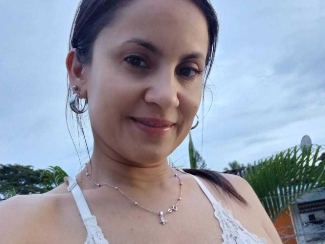 SolaraDubois with brown hair in a ponytail, wearing white lace top, hoop earrings, and a necklace with a small pendant, smiling subtly. Boobs visible, medium-sized. Outside, cloudy sky.