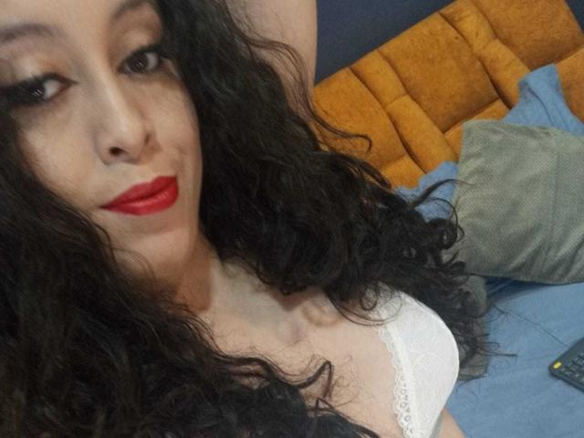 KendraSimons, avec ses cheveux noirs bouclés, son rouge à lèvres rouge et son soutien-gorge en dentelle blanche, est allongée sur un lit avec une tête de lit jaune et un oreiller bleu, et regarde l'appareil photo avec un léger sourire. Ses seins sont visibles..