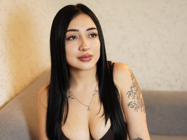MagicBeautyHot con lunghi capelli neri, occhi marroni, labbra lucide, indossa una top scollata che mette in mostra il suo seno prosperoso, un tatuaggio sul braccio destro, seduta su un divano grigio..
