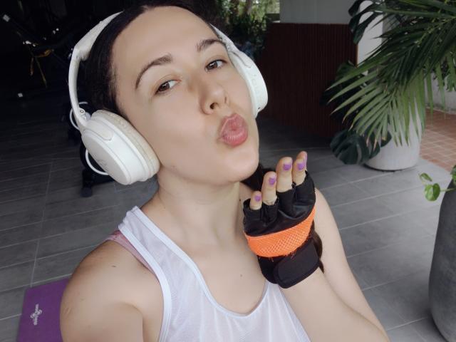 MabyLegrand with white headphones, pouty lips, purple nails, black and orange fingerless glove, white tank top, and dark hair, showing off small tits in a selfie.