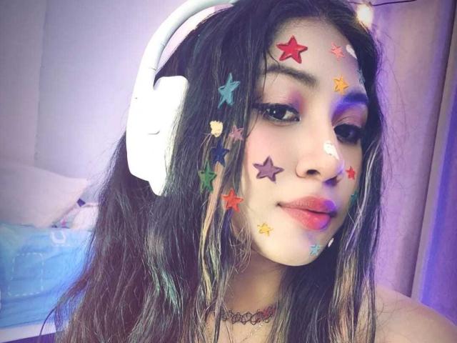 SiaraBonny with star face paint, colorful eyeshadow, white headphones, long dark hair, red lipstick, and a choker, looking cute and nerdy.