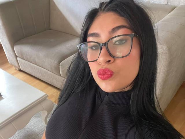 NinaRouse with black hair, glasses, and red lipstick, pouting her plump lips, wearing a black turtleneck, in a selfie, showing her big, round boobs in the foreground.