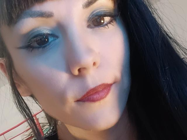 Das Gesicht von OrianaLaFrancaise mit blauem Lidschatten, schwarzen Eyeliner-Schwung, rotem Lippenstift und schwarzen Haaren im Seitenprofil. Ihre Haut ist blass, und sie hat ein leichtes Lächeln..