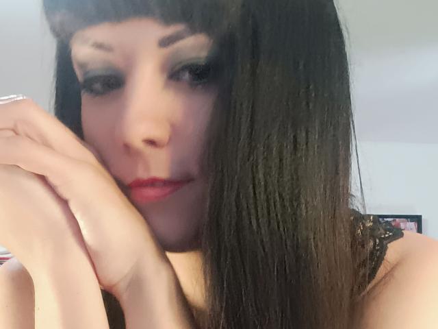 OrianaLaFrancaise, con capelli castano scuro e lisci, un pesante trucco occhi nero, rossetto rosso e pelle chiara, tiene le mani vicino al viso, guardando in basso con un sorriso appena accennato. Il seno è parzialmente visibile..
