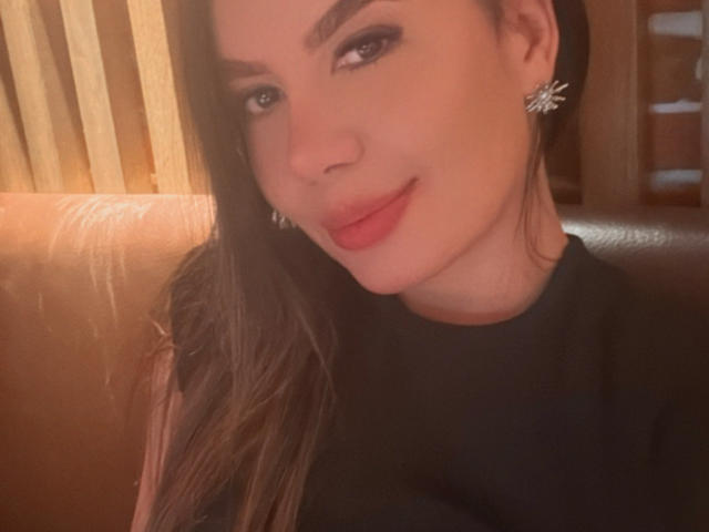 Valentina Eliott, con i suoi lunghi capelli castani, la camicia nera, il rossetto rosso e i brillanti orecchini, sorride dolcemente alla fotocamera, mostrando il suo viso liscio, carino e il leggero sorriso..