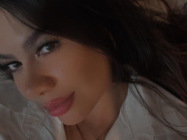 El rostro de Valentina Eliott, ojos marrón oscuro, cabello negro largo, labios rosados, ligera sonrisa, camisa blanca, luz suave, primer plano, maquillaje sutil, linda, sexy, atractiva, tono de piel cálido..