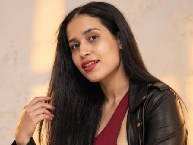 KinkyIris with long black hair, red lipstick, and brown eyes, wearing a black leather jacket over a red dress, slightly smiling, holding hair with her right hand.